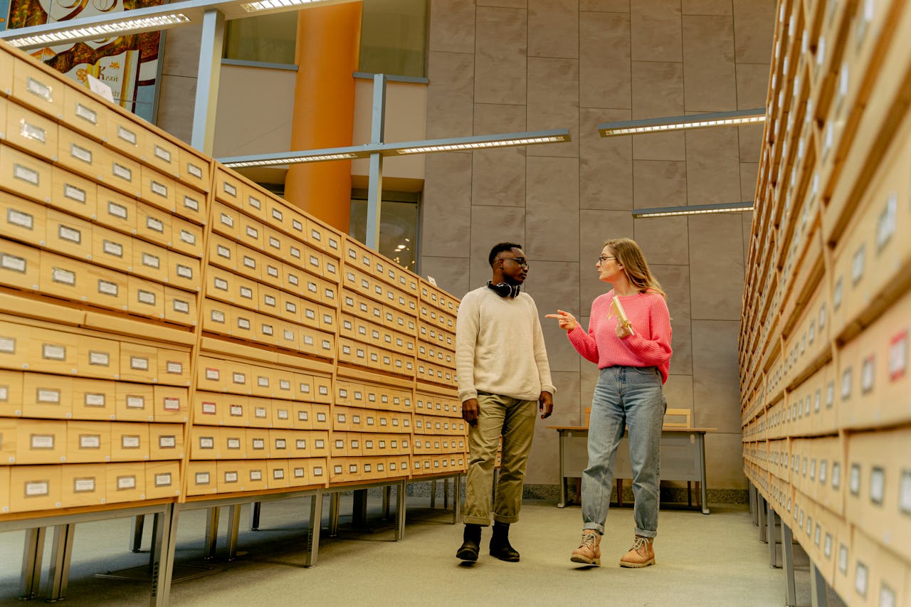 services-img Two adults conversing in a library archive aisle surrounded by wooden drawers and overhead lights.